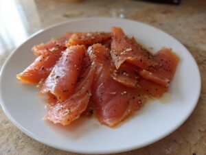 Close-up of thinly sliced smoked salmon seasoned with cracked pepper and sesame seeds on a white plate.
