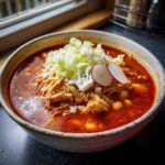 A rustic bowl filled with rich red chicken pozole, topped with shredded chicken, cabbage, and radish slices.