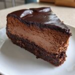A decadent slice of chocolate cheesecake with a thick, dark chocolate ganache topping, resting on a white plate.