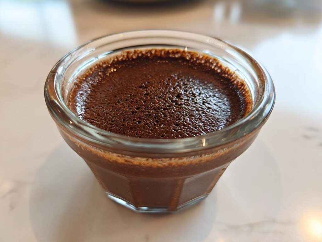Close-up of rich, dark chocolate mousse with a slightly bubbly top, served in a small, clear glass ramekin.