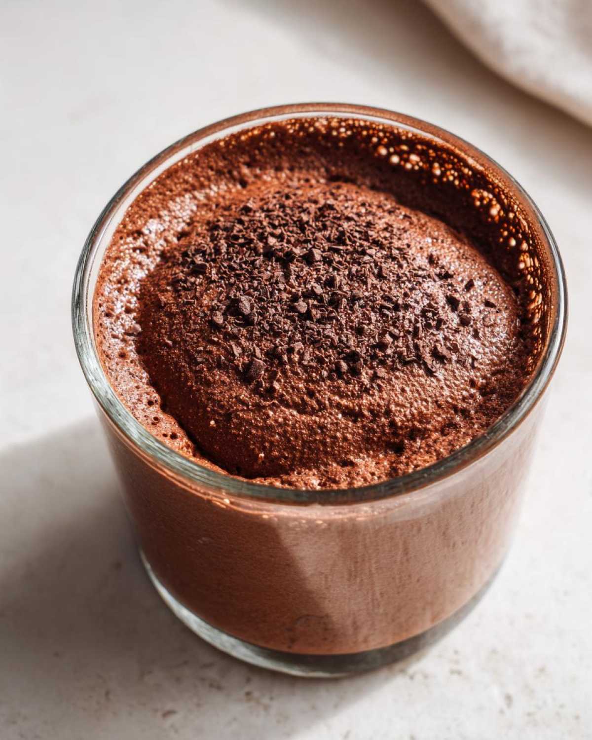 A close-up, overhead view of rich chocolate mousse topped with dark chocolate shavings served in a clear glass.