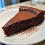A decadent slice of flourless chocolate torte dusted heavily with cocoa powder, served on a white plate.