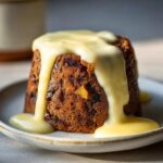A close-up of a traditional plum pudding covered in thick, pouring vanilla custard sauce.
