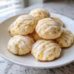 A pile of soft ricotta cookies drizzled with white lemon glaze and topped with zest on a white plate.