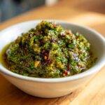 Close-up of a thick, vibrant green tomato salsa, chunky with herbs and chili flakes, served in a white bowl.