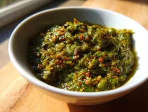 Close-up of thick, textured green tomato salsa with visible flecks of red spice, served in a small white bowl.