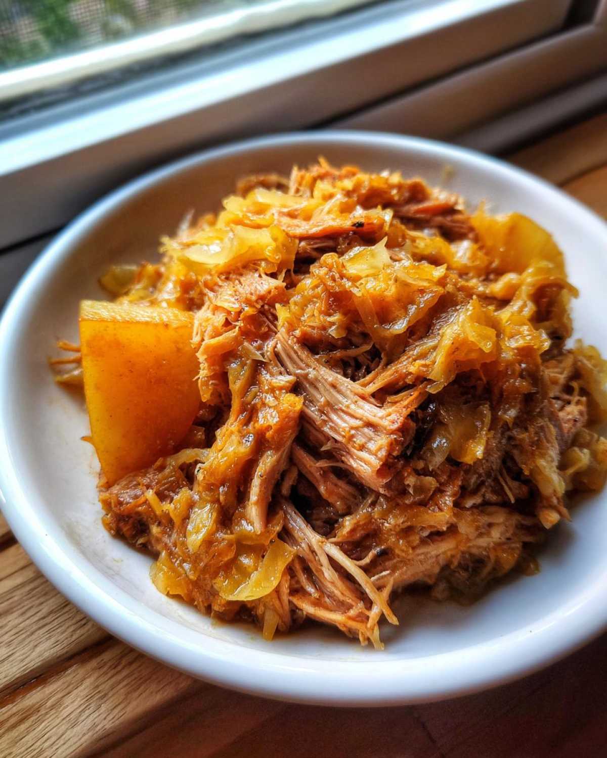 Close-up of a white bowl filled with tender, shredded pork and sauerkraut, served on a wooden surface.