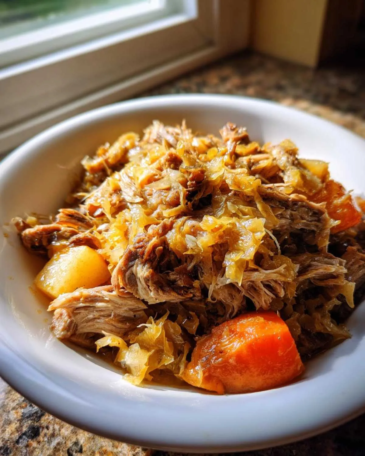 A close-up of shredded pork and sauerkraut mixed with chunks of carrots in a white bowl, featuring the pork and sauerkraut.