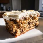 Close-up of a thick slice of chocolate chip cookie cake recipe topped with creamy white frosting.
