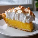 A close-up of a thick slice of bright yellow lemon pie topped with fluffy, torched meringue.
