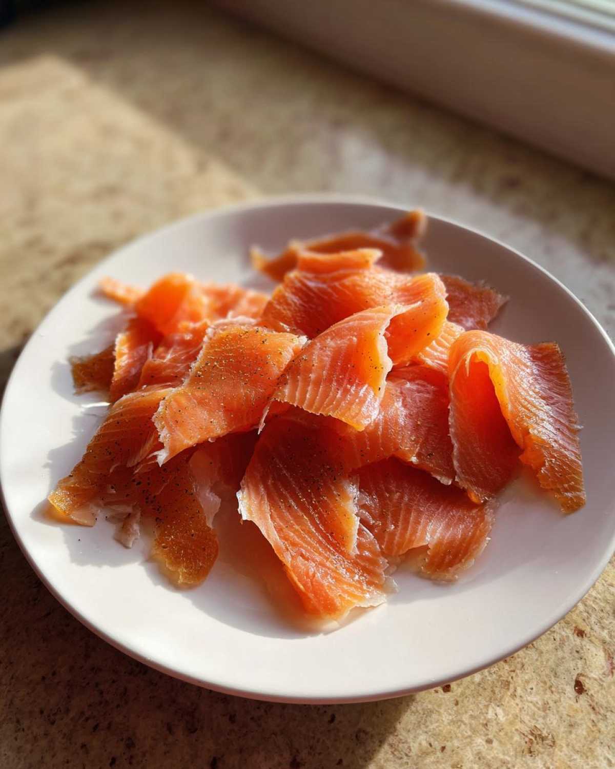 Close-up of thinly sliced, bright orange smoked salmon pieces seasoned with black pepper on a white plate.