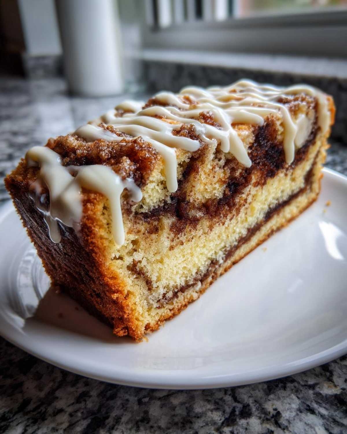 A thick slice of moist sour cream coffee cake featuring a cinnamon swirl and topped with a white vanilla glaze drizzle.