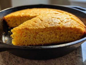 Close-up of a thick slice of moist sourdough cornbread with a golden crust, sitting in a black cast iron skillet.