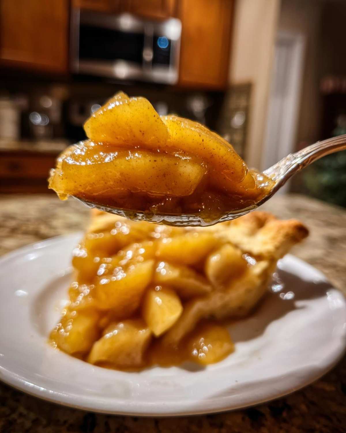 A spoonful of glistening, spiced apple pie filling held above a slice of pie on a white plate.
