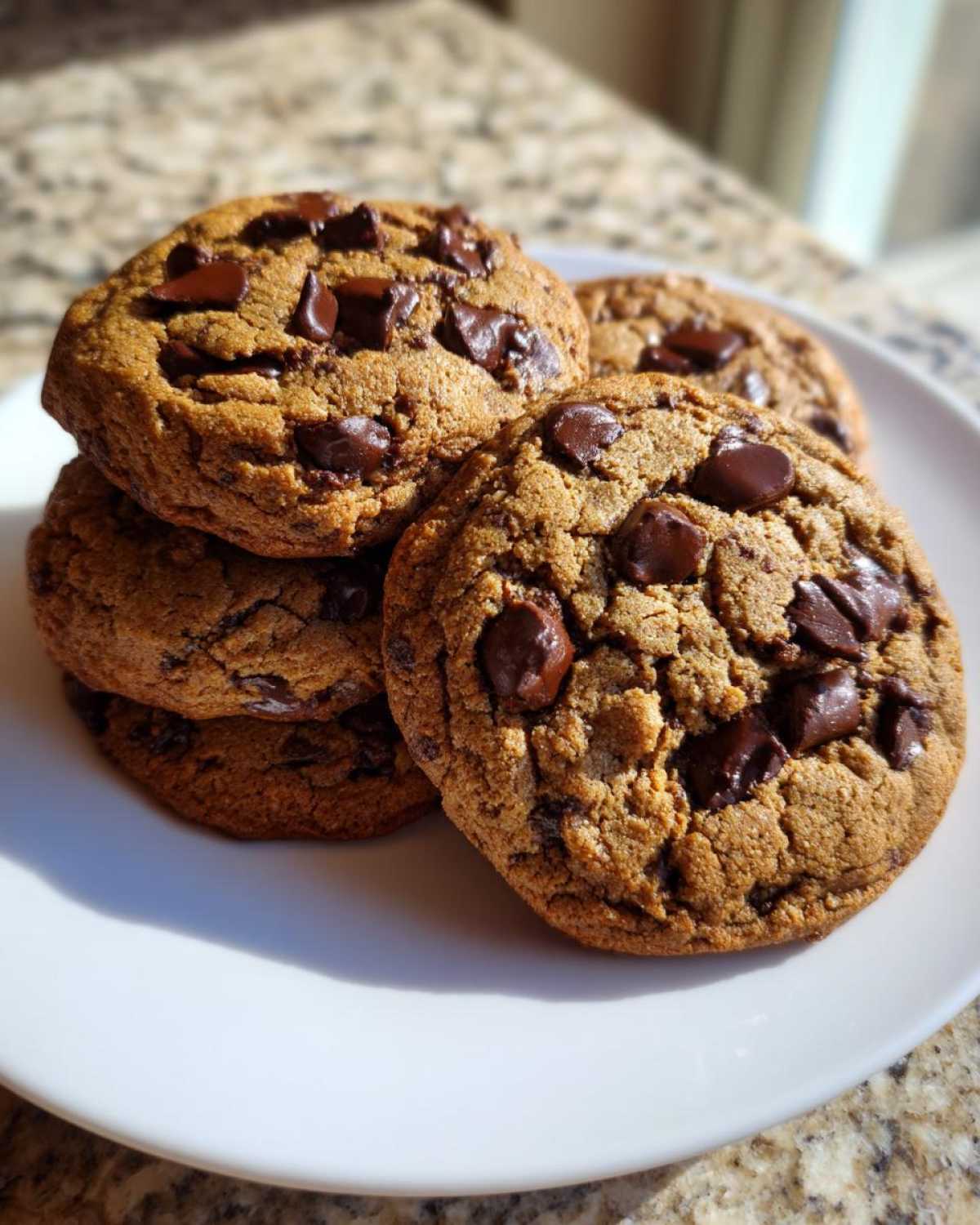 A close-up of several freshly baked vegan chocolate chip cookies stacked on a white plate, glistening in sunlight.