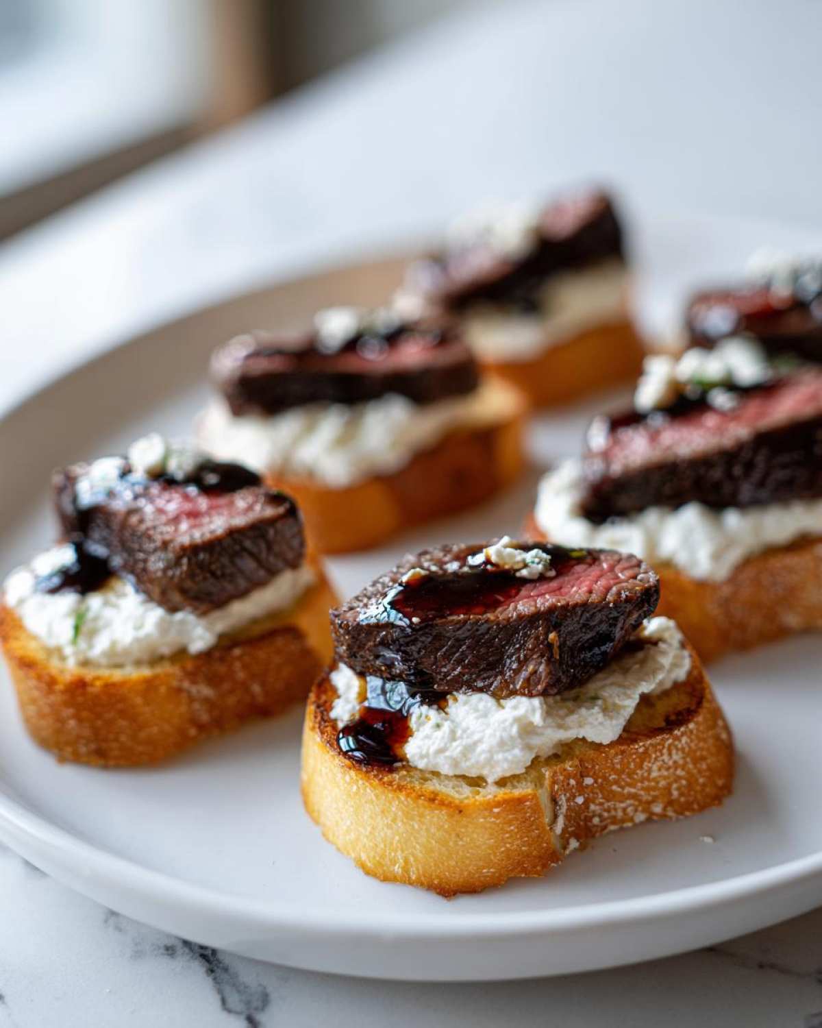 Close-up of several perfectly cooked steak crostini bites topped with creamy cheese and balsamic glaze.