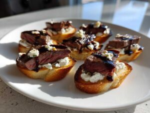Six pieces of steak crostini bites topped with rare steak, creamy cheese, and balsamic glaze, arranged on a white plate.