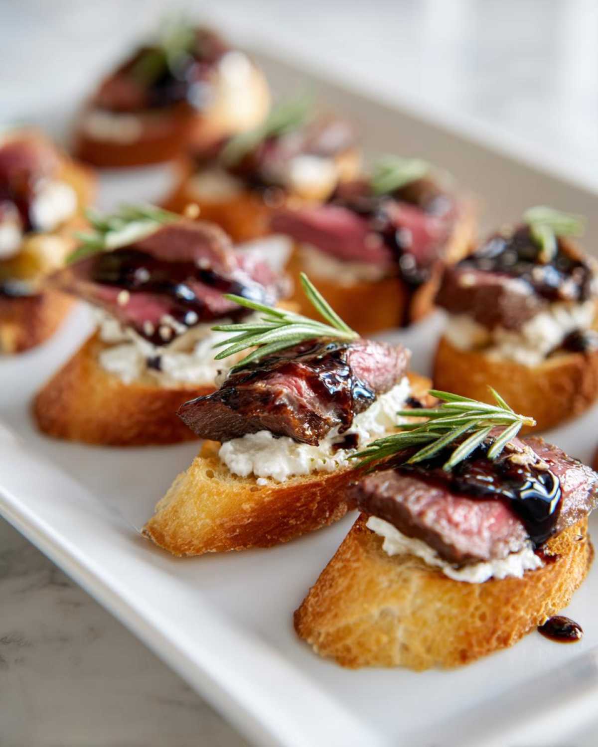 Close-up of several steak crostini bites topped with cheese, balsamic glaze, and rosemary on a white platter.
