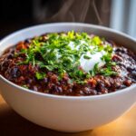 A close-up of a steaming white bowl filled with rich, dark black bean chili, topped with sour cream and fresh cilantro.