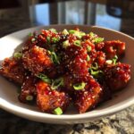 Close-up of sticky, glazed gochujang chicken pieces topped with sesame seeds and sliced green onions.
