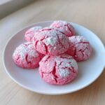 Five bright pink strawberry cake mix cookies dusted heavily with white powdered sugar on a white plate.