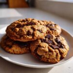A close-up of four freshly baked vegan chocolate chip cookies stacked on a white plate.