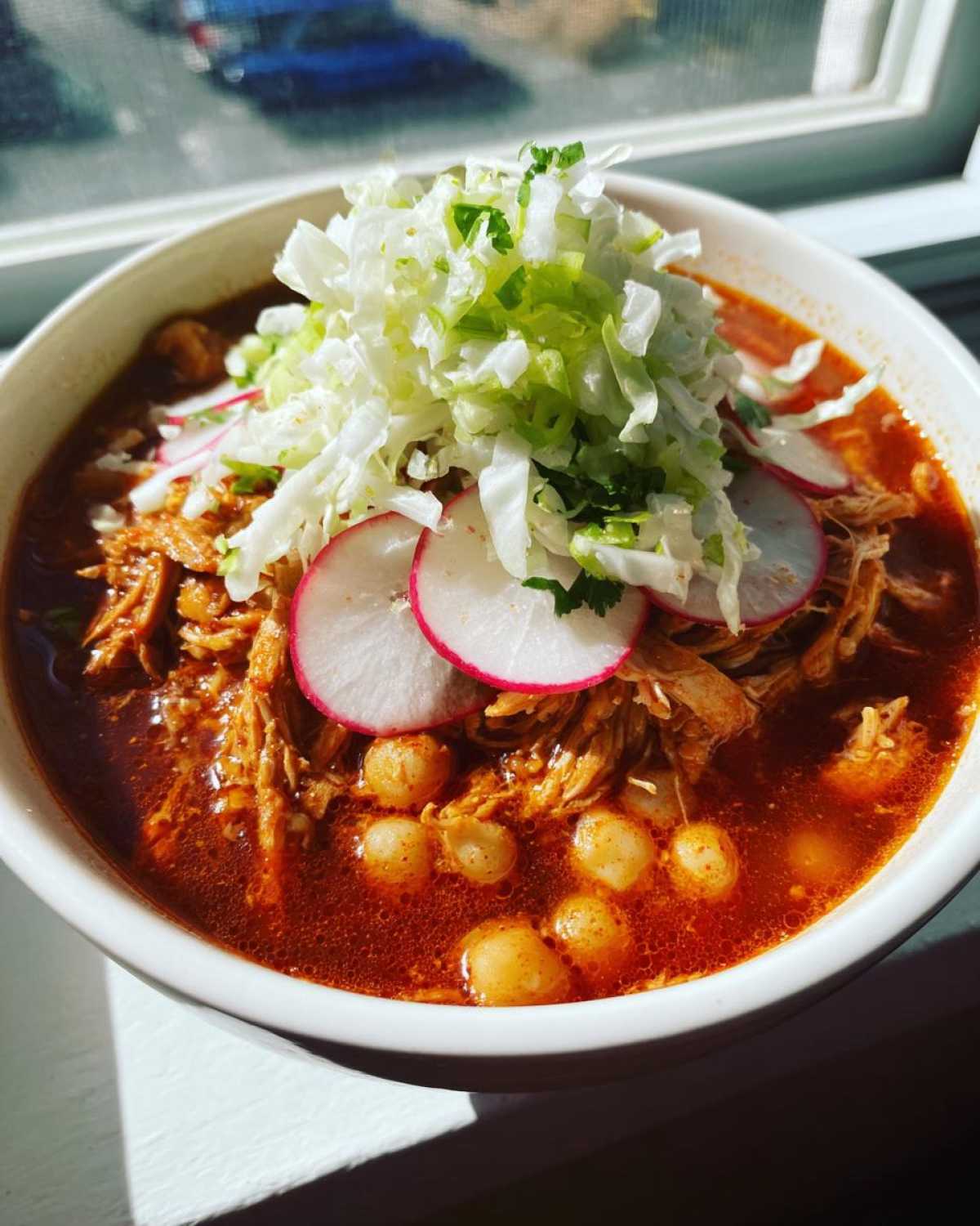 A vibrant bowl of red chicken pozole featuring shredded chicken, hominy, and topped generously with shredded lettuce and radish slices.