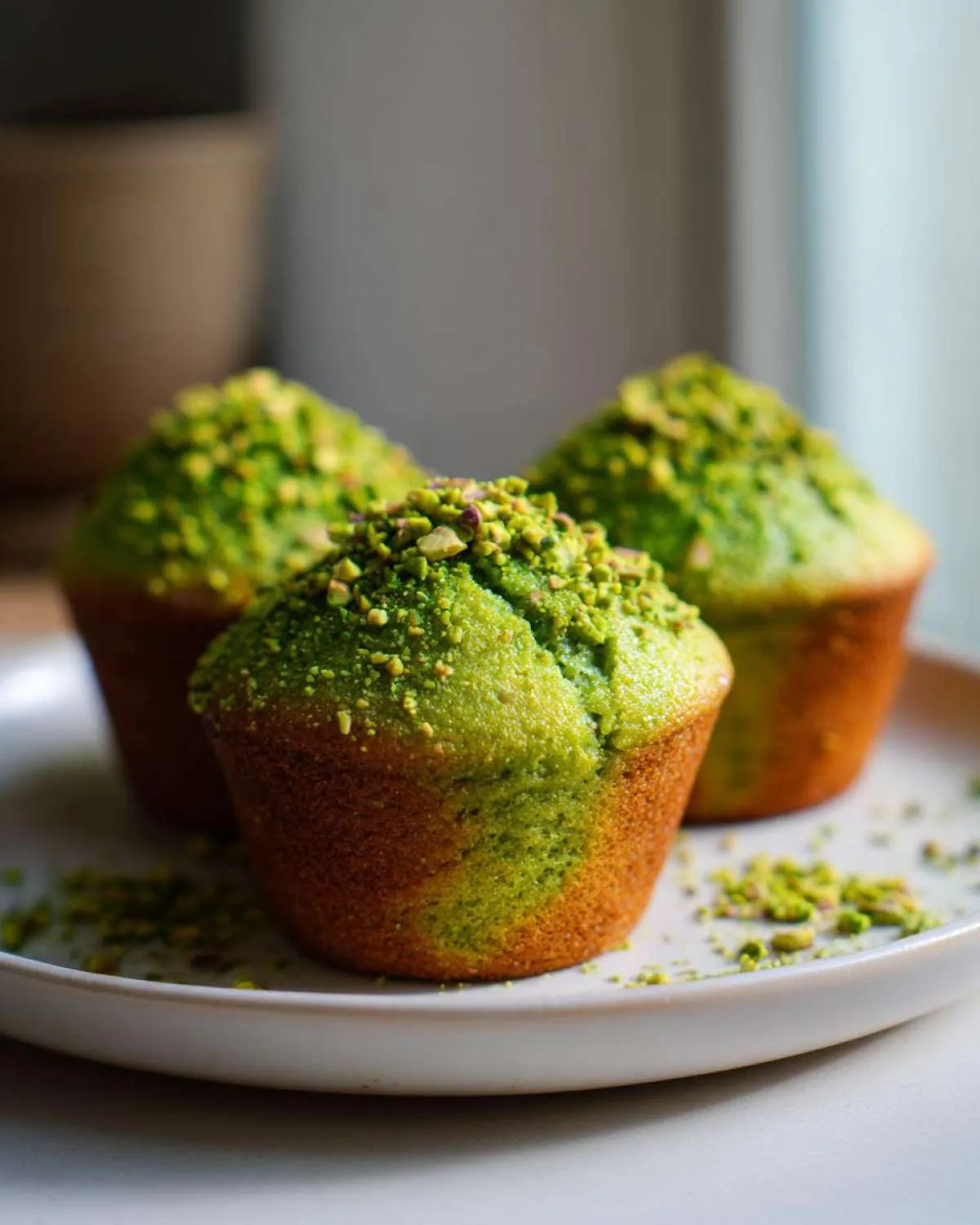 Three bright green pistachio muffins topped with chopped nuts, sitting on a light plate.