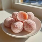 Close-up of several pink watermelon mochi balls dusted with white powder on a small white plate.
