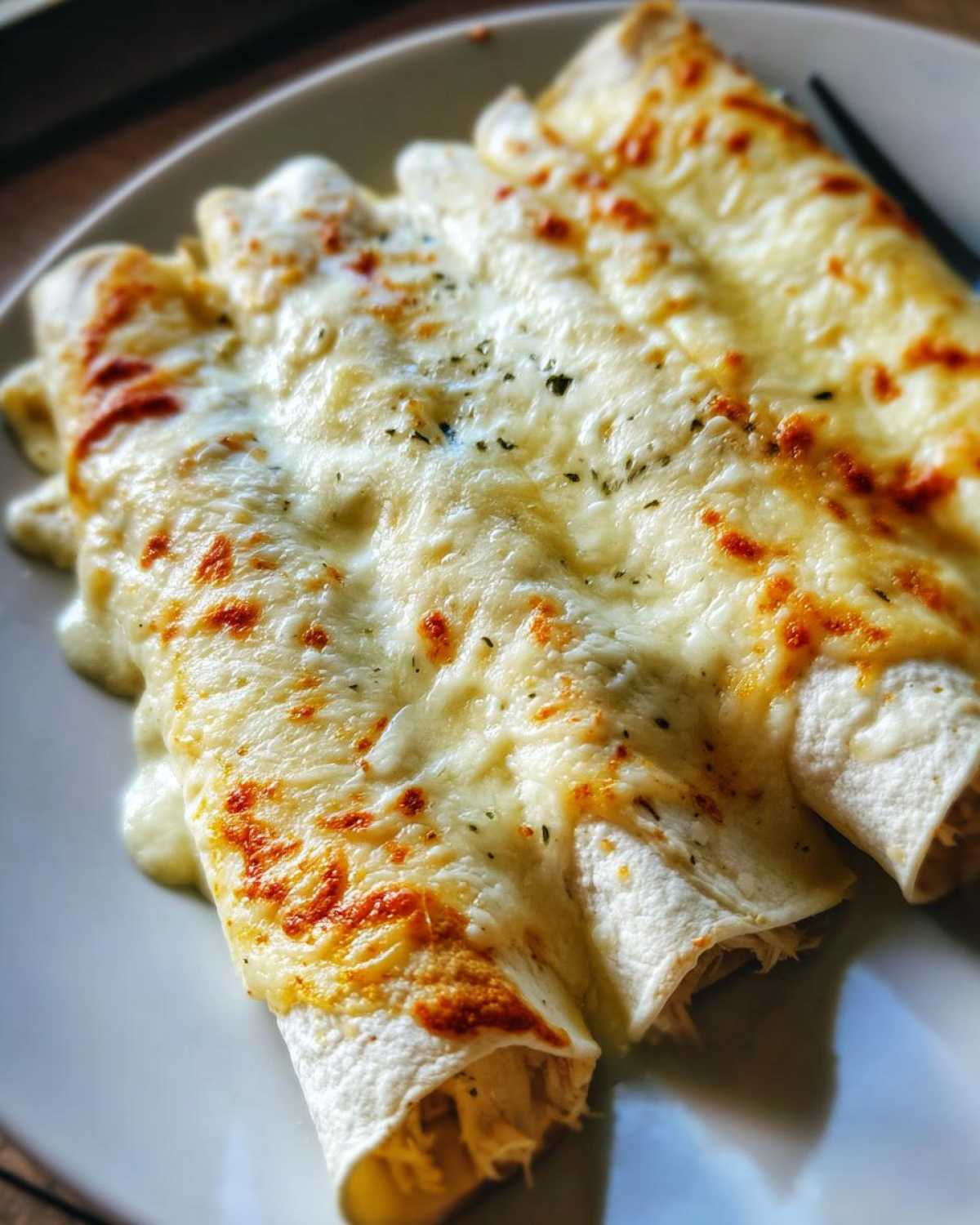 Close-up of three rolled white chicken enchiladas covered in melted, slightly browned cheese and sprinkled with herbs.