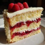 A close-up slice of white chocolate raspberry cake featuring layers of yellow sponge, white frosting, and bright red raspberry filling, topped with fresh raspberries.