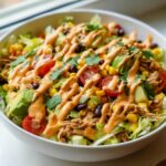 A vibrant bowl of southwest salad featuring shredded chicken, avocado, black beans, corn, tomatoes, and a creamy orange dressing drizzle.