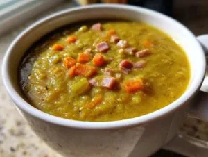 A close-up shot of thick, green pea soup topped with diced ham and carrots in a white mug.