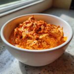 Close-up of a creamy, orange-colored chili cheese dip topped with spices in a white bowl.