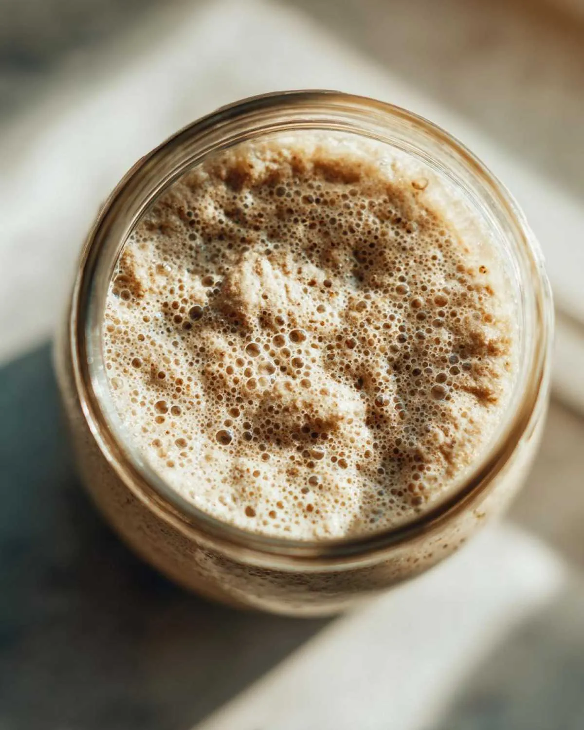 Overhead view of a bubbly, active sourdough starter in a glass jar, showing signs of fermentation.