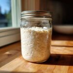 A glass jar filled with bubbly, active sourdough starter sitting on a sunlit wooden countertop.