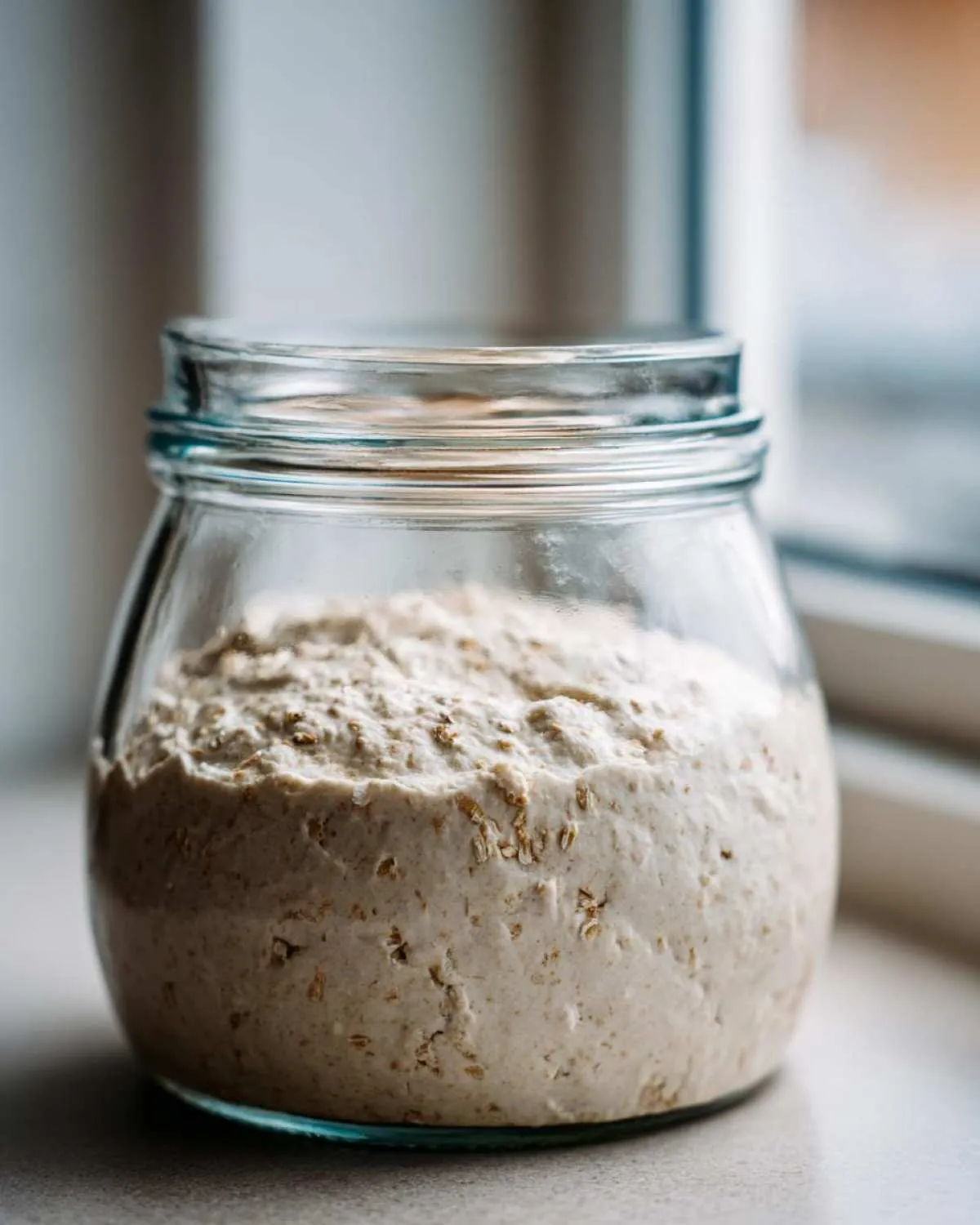 A close-up of an active sourdough starter, showing bubbles and whole grain flakes, resting in a clear glass jar.