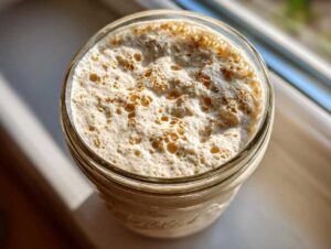 Overhead view of an active, bubbly sourdough starter in a glass jar, dusted with cinnamon or flour.