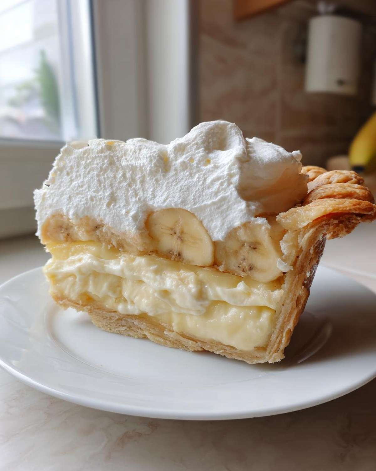 A tall slice of amazing banana cream pie showing layers of flaky crust, thick custard, banana slices, and whipped cream topping.