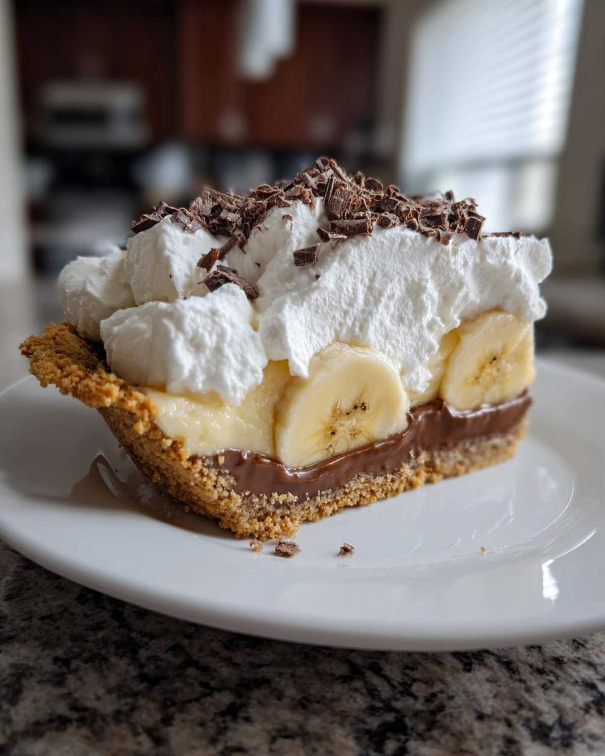 A close-up slice of rich banoffee pie showing layers of crust, chocolate, banana, whipped cream, and chocolate shavings.