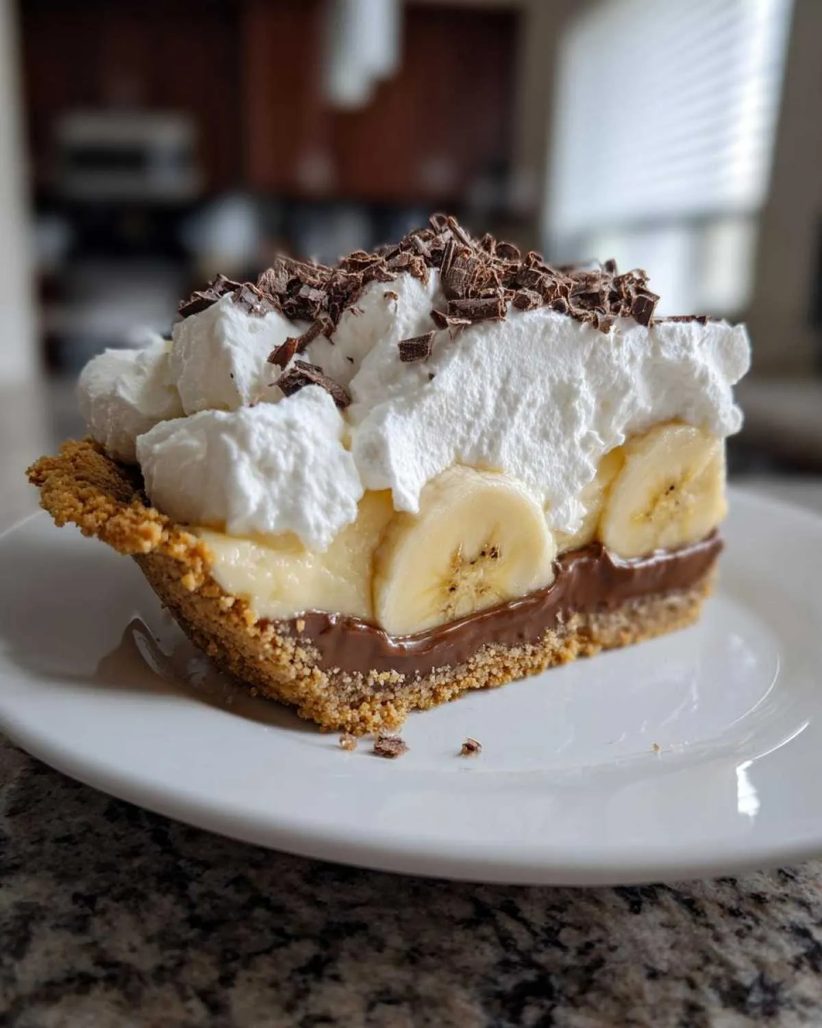 A close-up slice of rich banoffee pie showing layers of crust, chocolate, banana, whipped cream, and chocolate shavings.