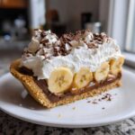 A decadent slice of banoffee pie featuring a graham cracker crust, layers of caramel and chocolate, sliced bananas, whipped cream, and chocolate shavings.
