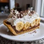 A decadent slice of banoffee pie featuring a graham cracker crust, layers of caramel and chocolate, sliced bananas, whipped cream, and chocolate shavings.