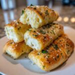 A stack of soft, golden-brown garlic breadsticks topped with melted butter, parsley, and Parmesan cheese.