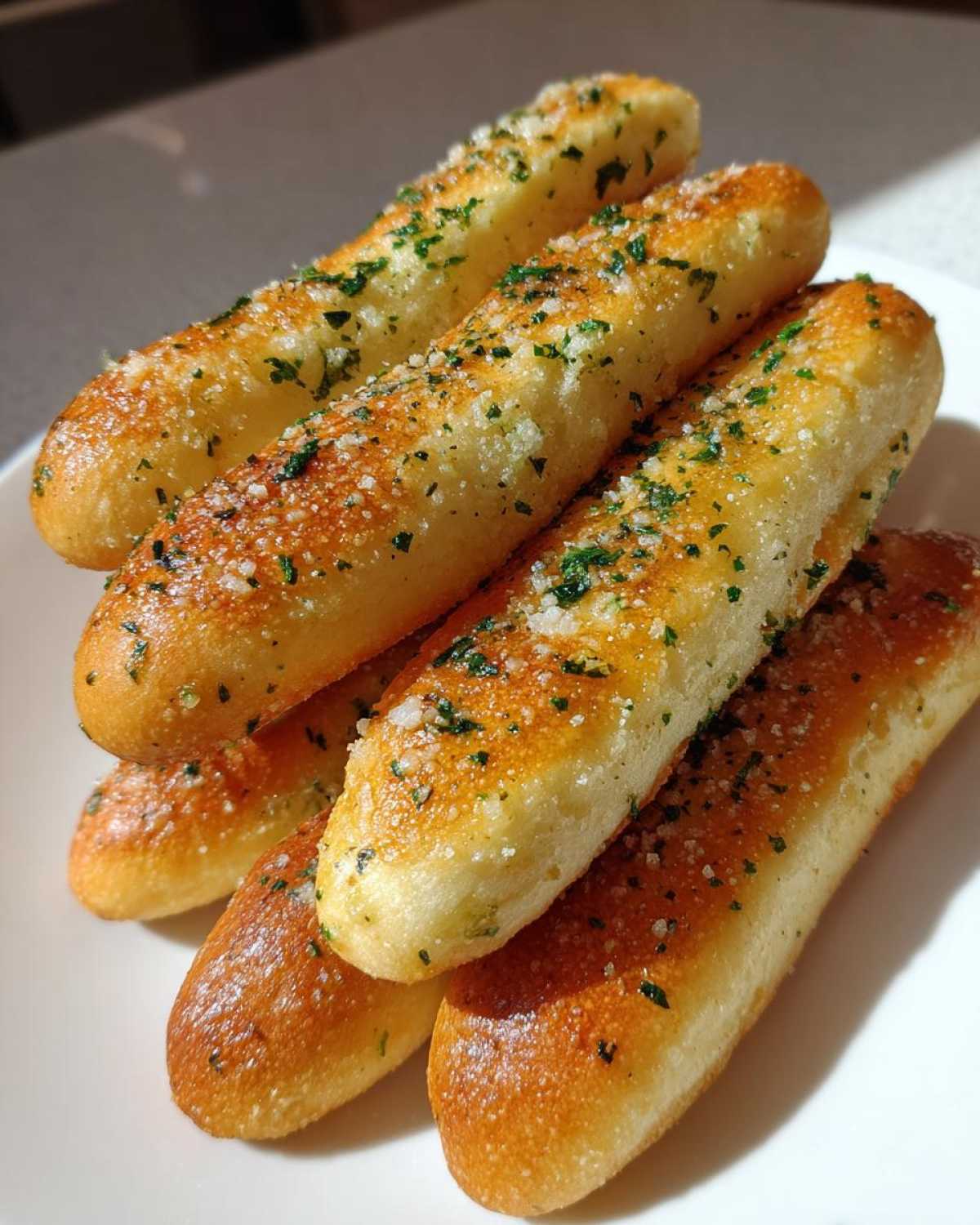 A stack of freshly baked, soft breadsticks seasoned with garlic, salt, and green parsley flakes.