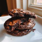 Close-up of three stacked, fudgy brownie cookies with shiny, cracked tops and visible melted chocolate chips.