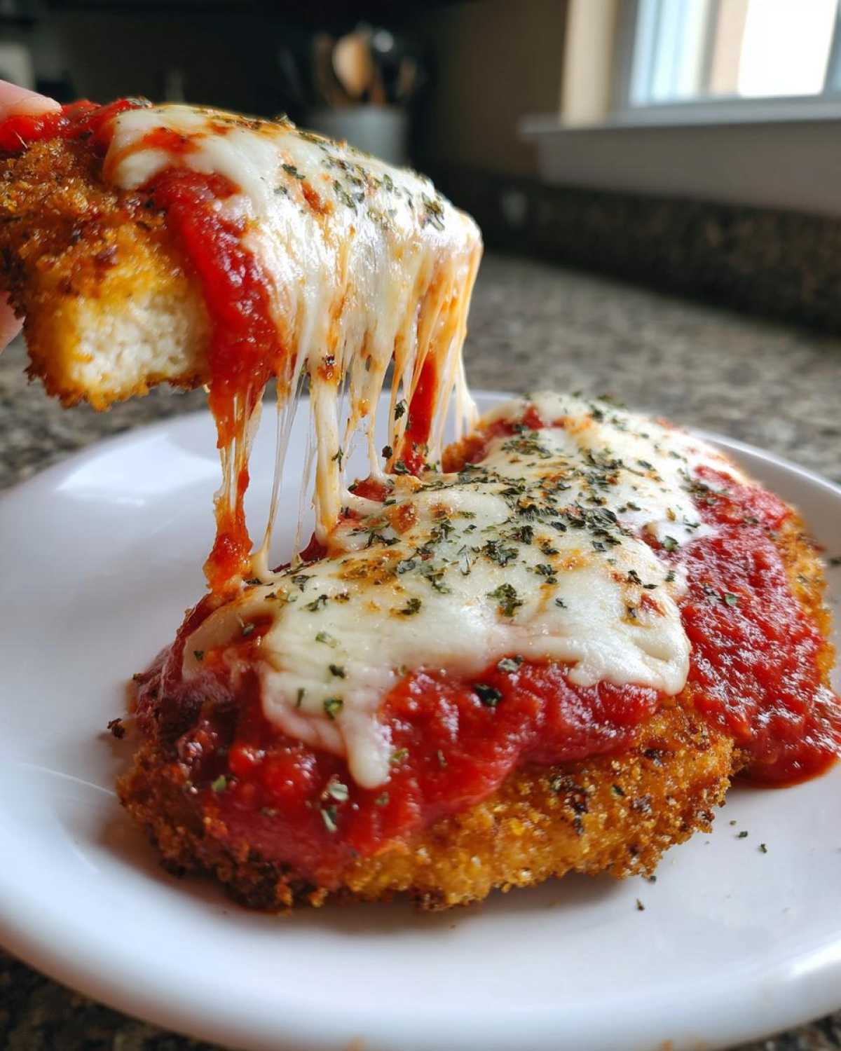 A piece of crispy chicken parmesan being lifted, showing a dramatic cheese pull over the saucy main dish.