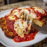 Close-up of a piece of crispy chicken parmesan being lifted, showing a dramatic, long cheese pull over marinara sauce.