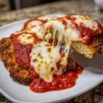 Close-up of a piece of crispy chicken parmesan being lifted, showing a dramatic, long cheese pull over marinara sauce.