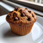 A close-up of a freshly baked chocolate chip muffin with melted chips on top, sitting on a white plate.